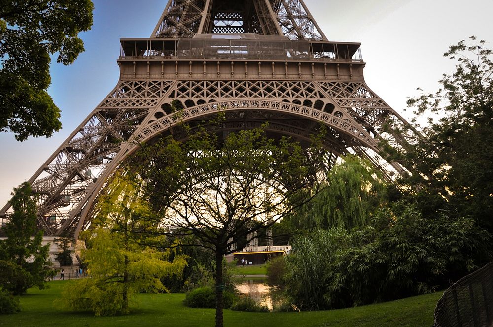 A close-up shot of the bottom of the Eiffel Tower with trees and water beneath it. Original public domain image from…