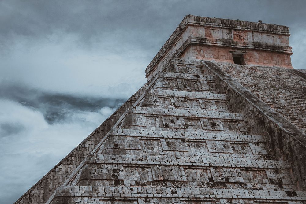 Looking up at an ancient stone pyramid under a cloudy sky in Chichén Itzá. Original public domain image from Wikimedia…