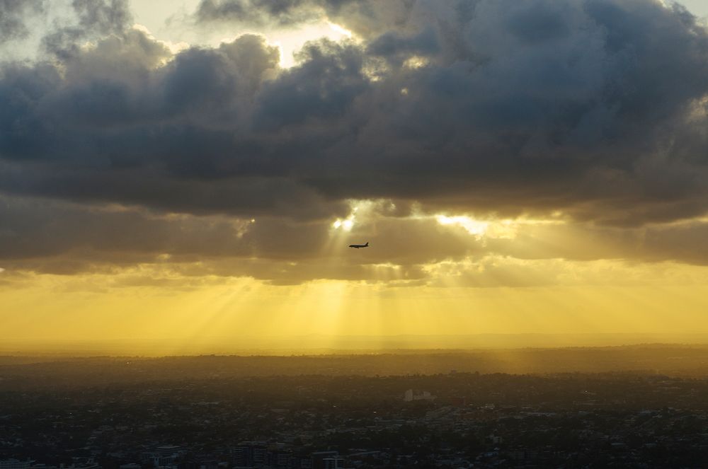 The silhouette of a passenger plane in the sky with sun breaking through heavy clouds above and a city below. Original…