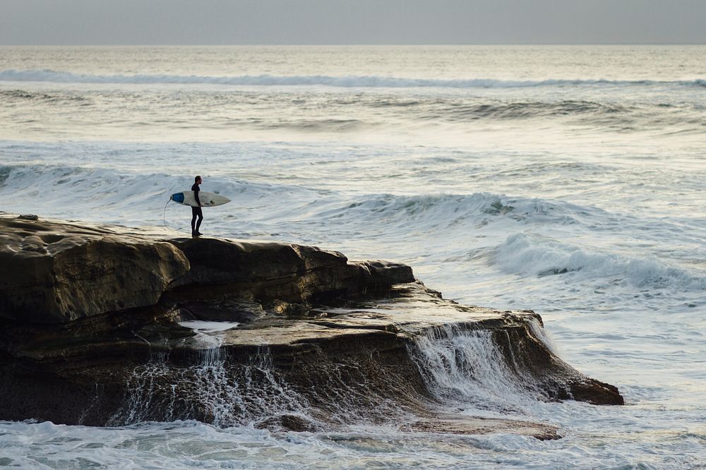 Surfer with a surfboard standing on a coastal rock watching rough ocean at Ocean Beach. Original public domain image from…