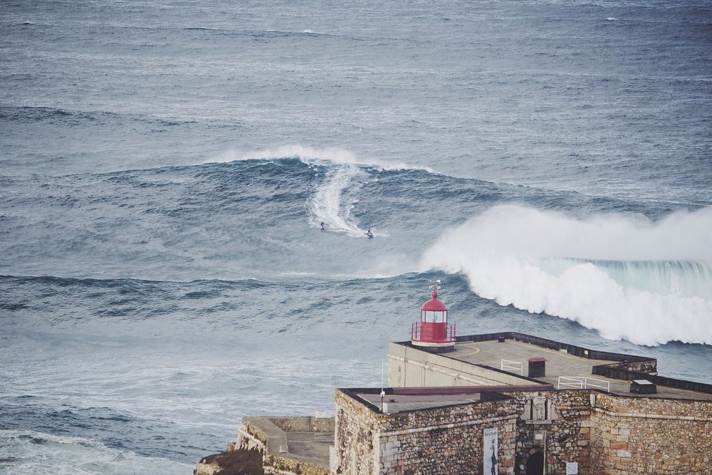 An ocean wave curling in the distance, in front of a coastal lighthouse at Nazaré. Original public domain image from…