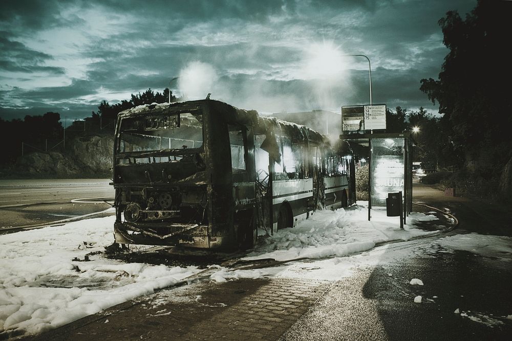 A burnt down bus on a patch of fire fighting foam next to a bus stop. Original public domain image from Wikimedia Commons