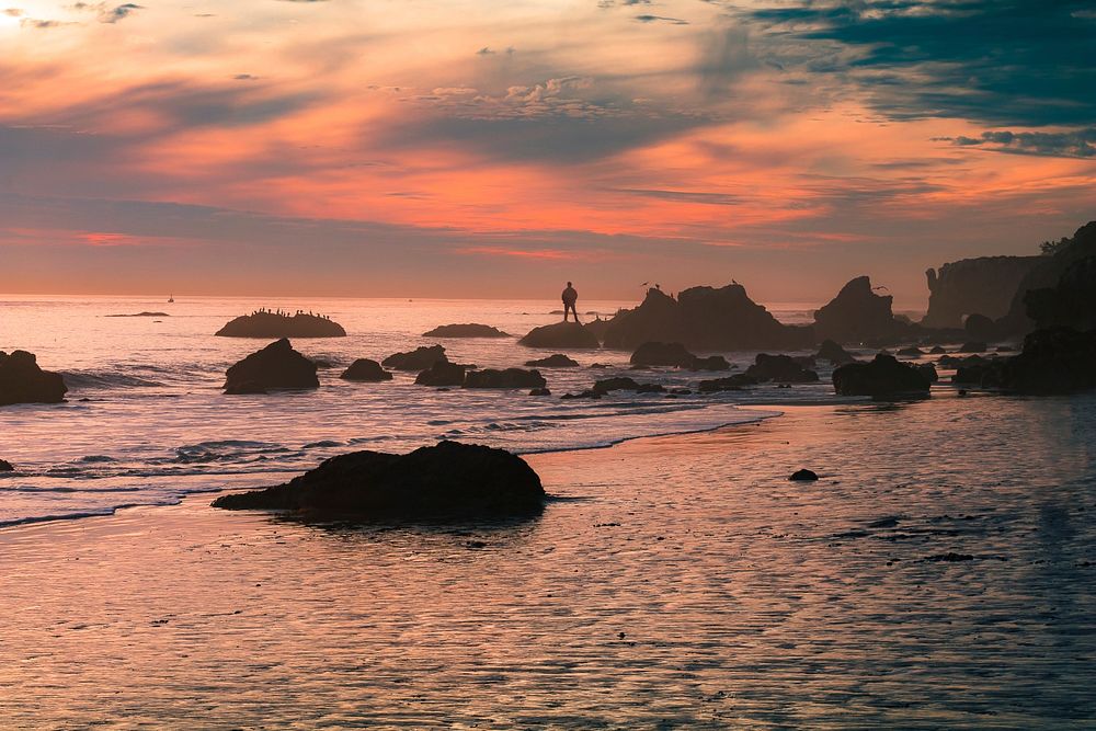 A pastel sunset covers the shores of El Matador State Beach as a man stands over the rocks. Original public domain image…