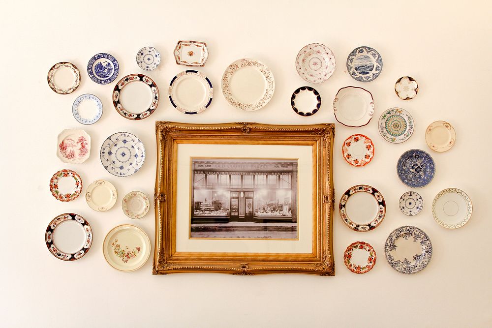 Photo in a wooden frame surrounded by colorful decorative plates all hanging on a white wall. Original public domain image…