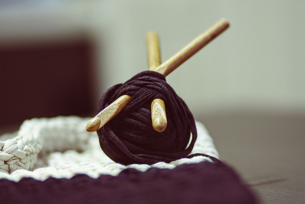 A roll of yarn with wooden knitting needles pierced through next to a weaved fabric. Original public domain image from…