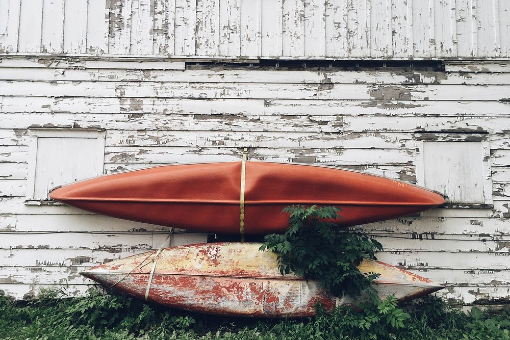 Two used kayaks leaning on an old barn wall with peeling white paint and unkept grass.. Original public domain image from…