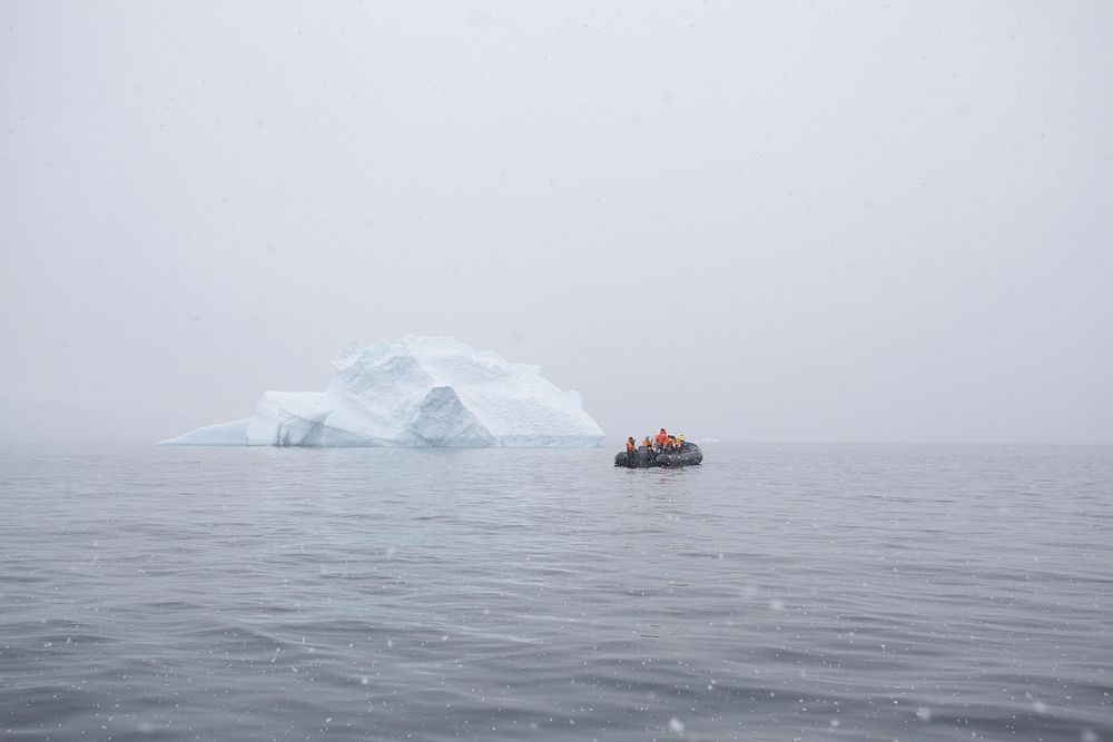 A group of people in a raft in the middle of the ocean next to a small iceberg with fog surrounding the area.. Original…