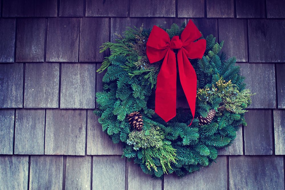 A Christmas wreath with a red bow and pinecones hanging on the side of a shingled house. Original public domain image from…