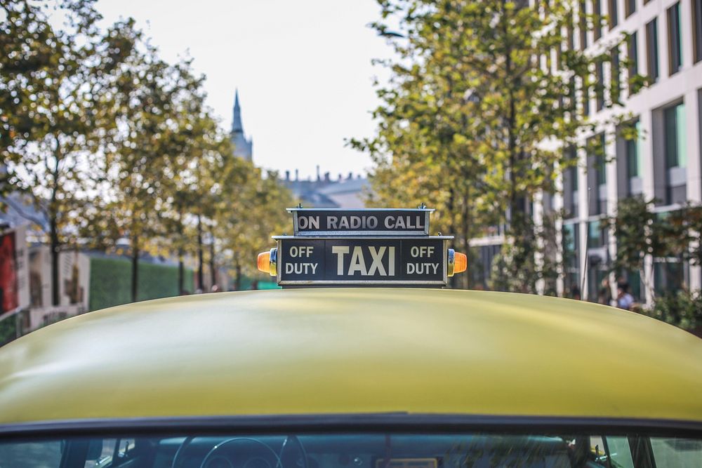 Close up shot of top of yellow London taxi cab sign on road with trees. Original public domain image from Wikimedia Commons