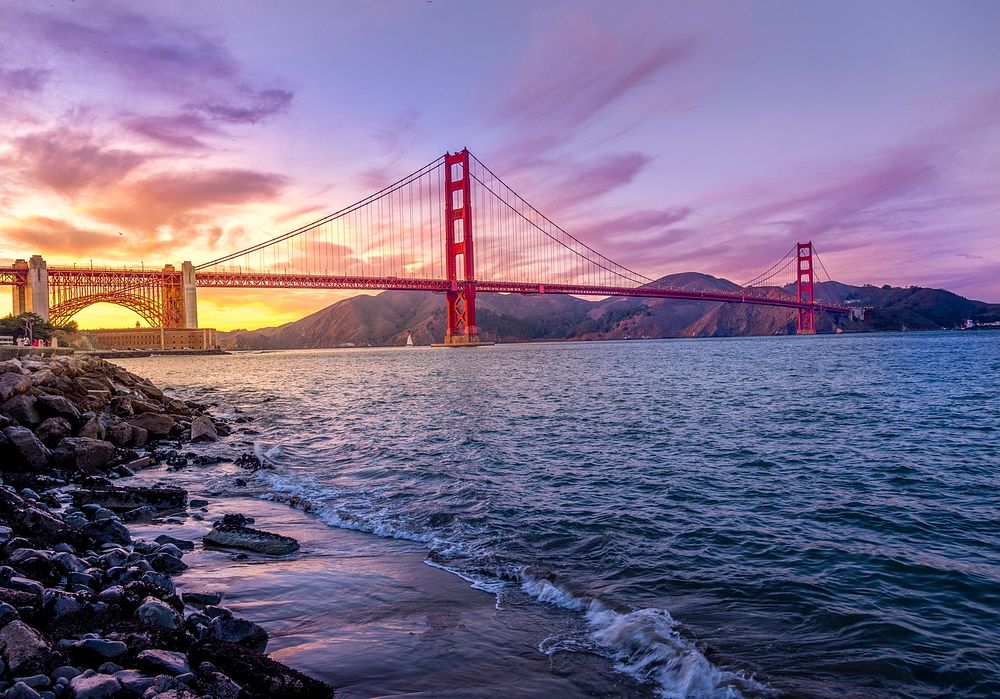 A purple and yellow sunset behind Golden Gate Bridge in San Fransisco, with waves breaking on the shoreline.. Original…