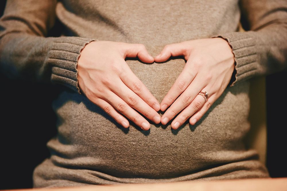 A pregnant woman in a sweater forming a heart with her hands on her belly. Original public domain image from Wikimedia…