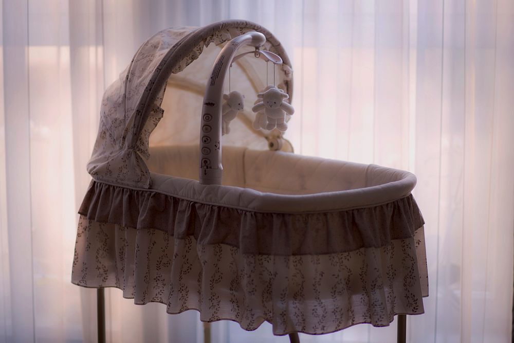 Soft hued bassinet with baby mobile hanging over the opening in front of light filled white curtains. Original public domain…