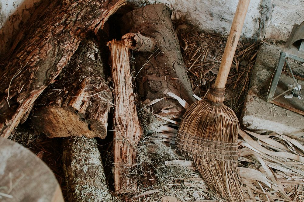 A broom leaning against a wall next to a disorderly heap of firewood. Original public domain image from Wikimedia Commons