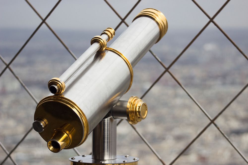 Sleek telescope peers through a chainlink fence over the city. Original public domain image from Wikimedia Commons