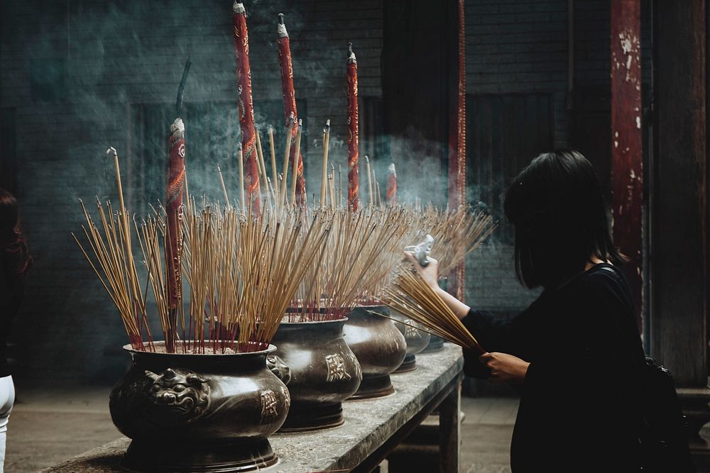 A woman places joss sticks in urns with joss sticks of various sizes in the temple.. Original public domain image from…