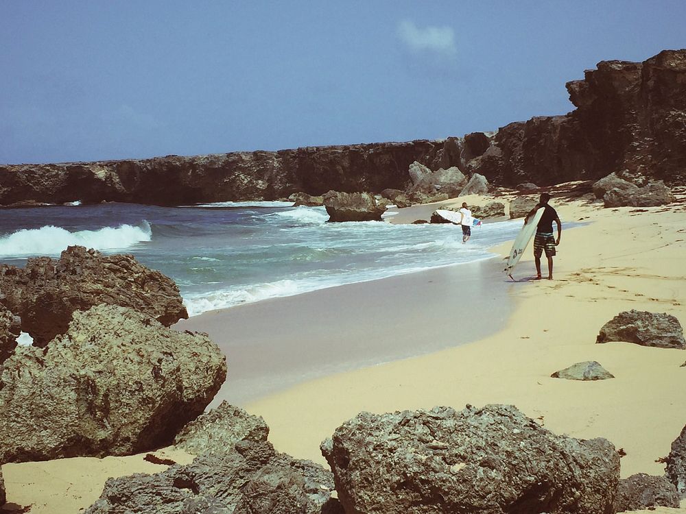Two surfers on the sand beach in bay among rocky cliffs. Original public domain image from Wikimedia Commons