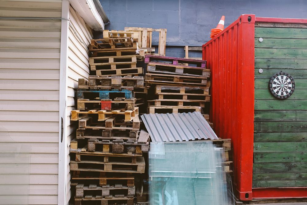 Pallets stacked by corrugated steel and garbage beside shipment containers and a dartboard. Original public domain image…