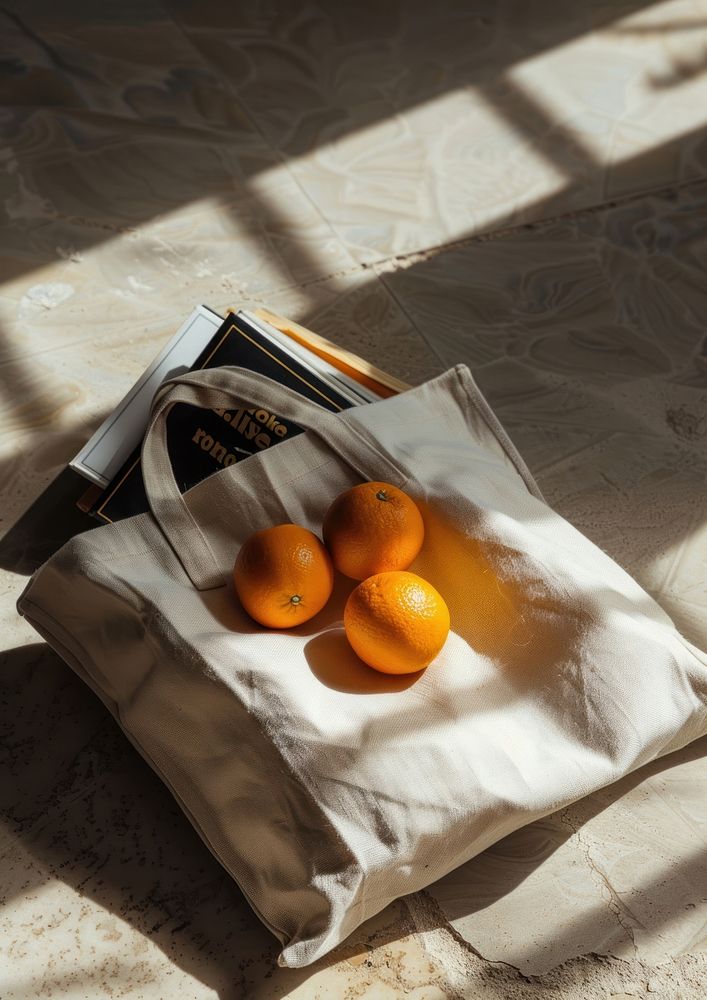 An open tote bag lying on the ground filled with book and two oranges furniture produce cushion.