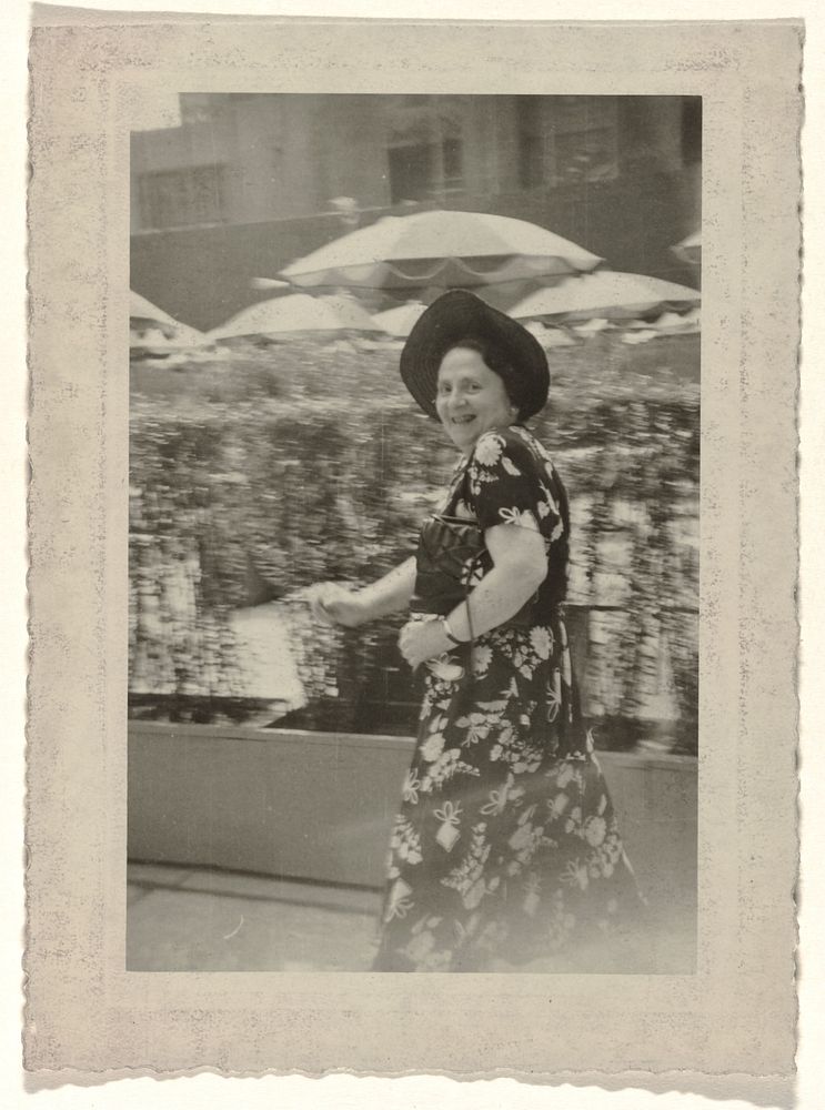 Tante Rosel in jurk met versierd motief en hoed op haar hoofd, lopende voor een struikenhaag met parasols (1950) by anonymous