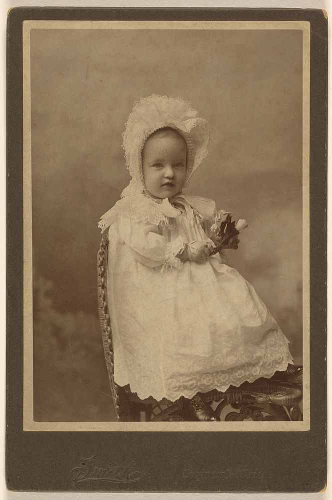 Unidentified baby wearing a bonnet, holding a flower, seated by Smith