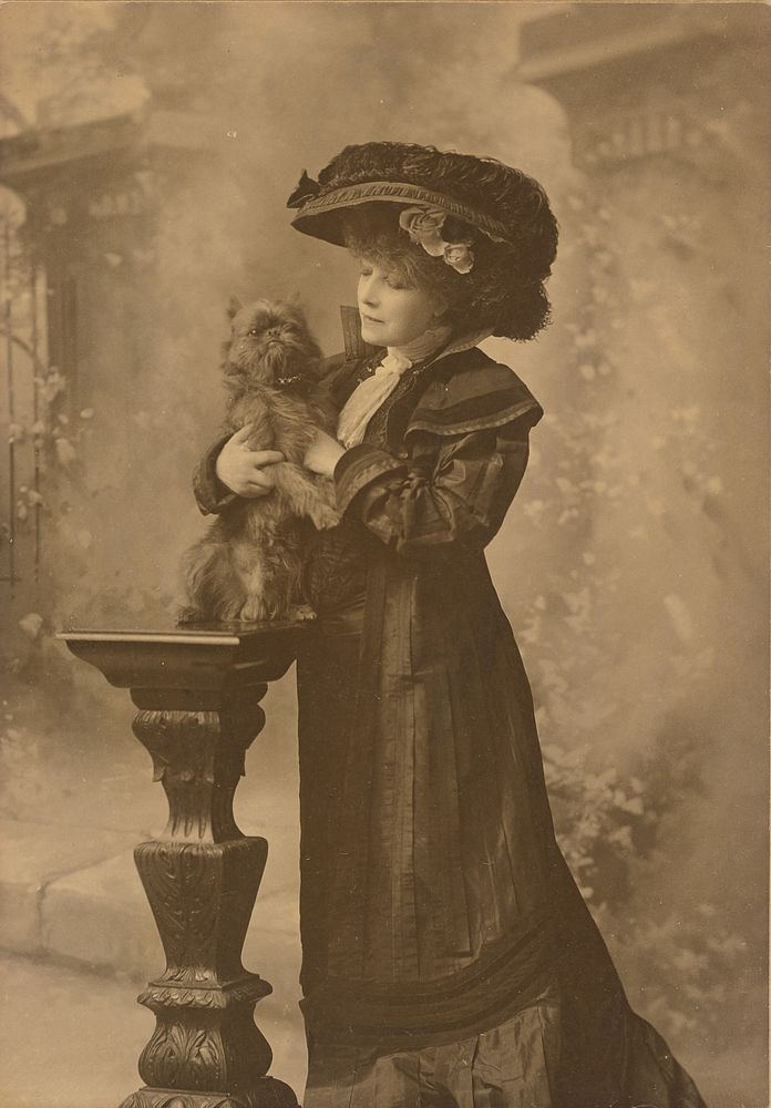 Sarah Bernhardt standing in a studio setting, holding a little black dog on an ornate, carved wooden pedestal by Theodore C…