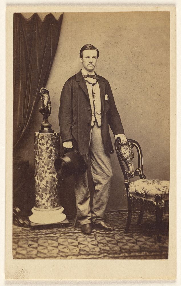 Unidentified man with moustache standing, holding top hat in one hand, the other hand on chair back by F W Baker