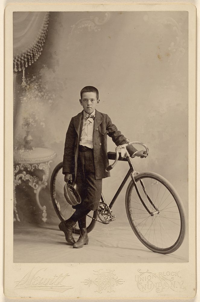 Unidentified young boy with cap in hand, standing with bicycle, in a studio setting by Hewitt