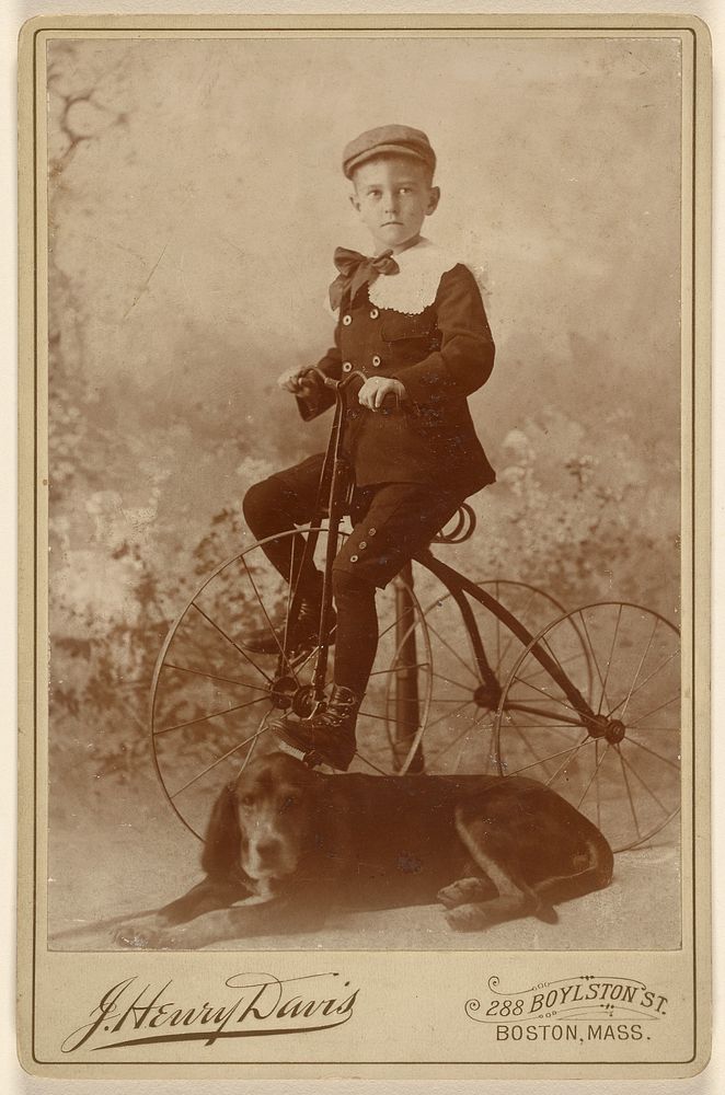 Unidentified little boy wearing a cap, mounted on an old-style three-wheel bicycle, with a dog in the foreground by J Henry…