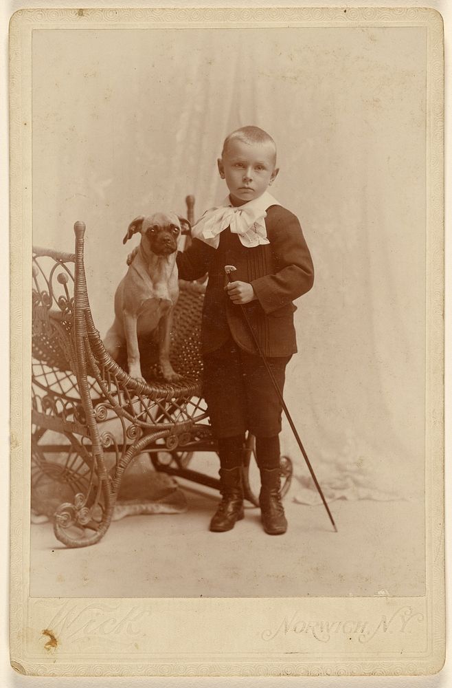 Little boy standing, holding a cane, with a little dog in a wicker chair by G Wick