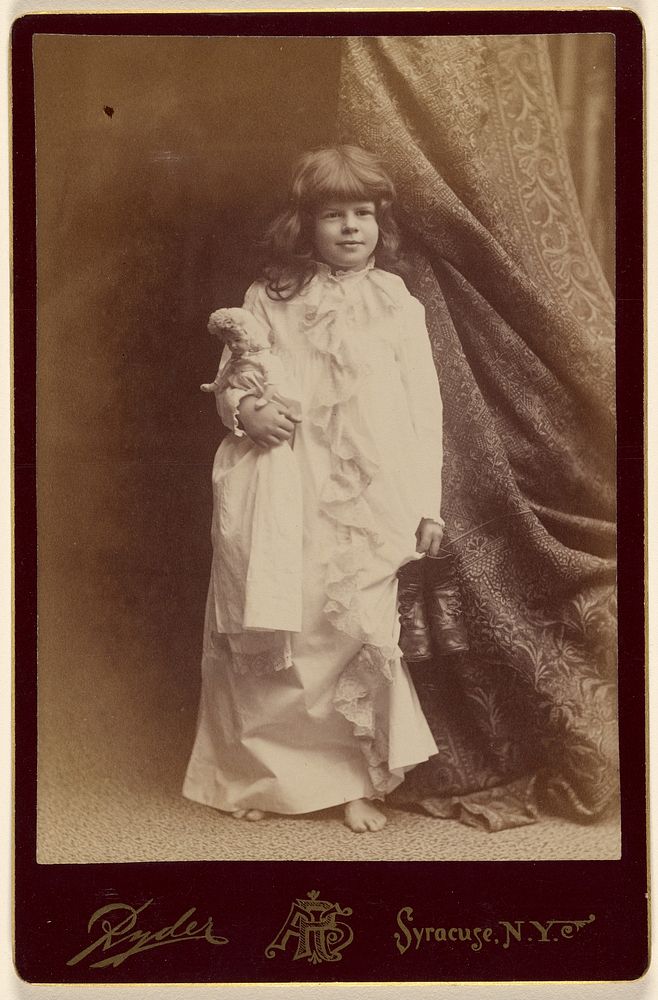 Ethel Butter in white dressing gown, holding pair of shoes and a doll, standing by Philip S Ryder