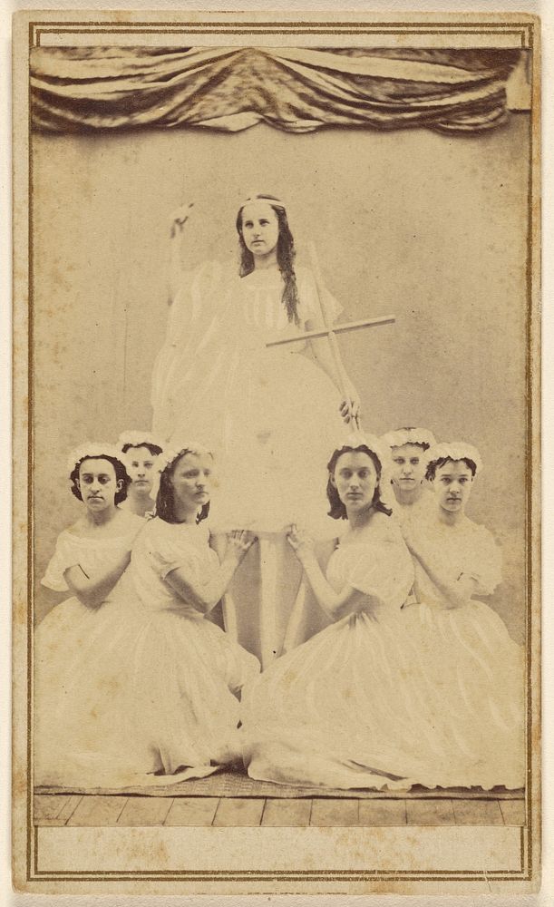 Female group portrait, six in white gowns kneeling, one standing holding a cross