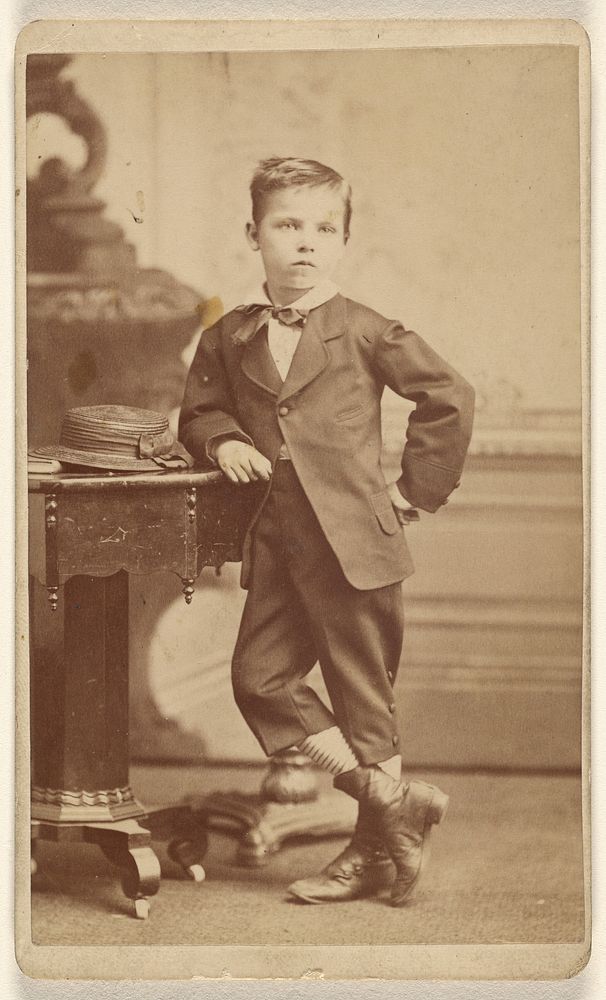 Young boy in suit standing and posed near a table with a hat on top by McClain