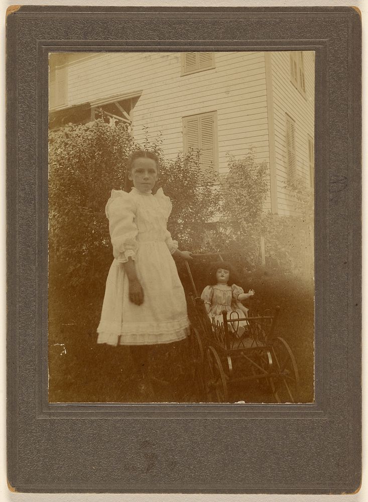 Unidentified little girl in white dress standing next to a baby carriage with a doll inside