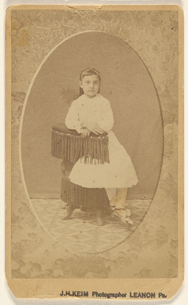 Unidentified little girl posed on chair with tassels, printed in quasi-oval style by J H Keim