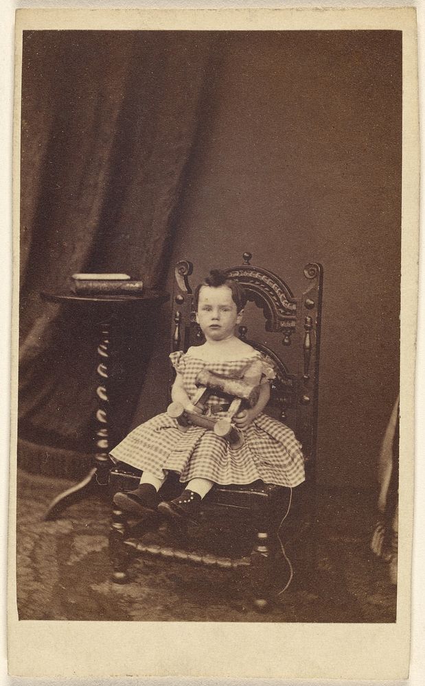 Unidentified little girl holding a toy wooden horse, seated