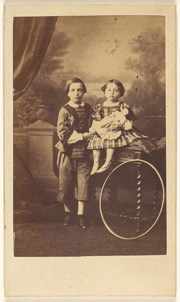 Unidentified little boy standing next to an unidentified little girl seated on a table, a large hoop resting on a table leg…