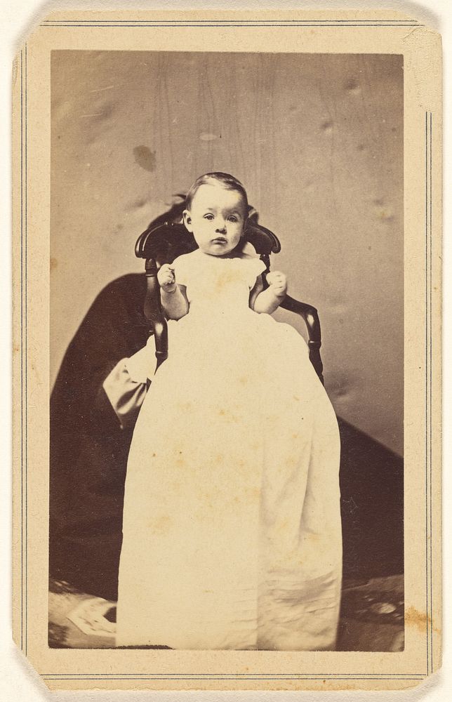 Unidentified baby wearing a long dressing gown, with unidentified figure behind holding up baby by J H Grotecloss