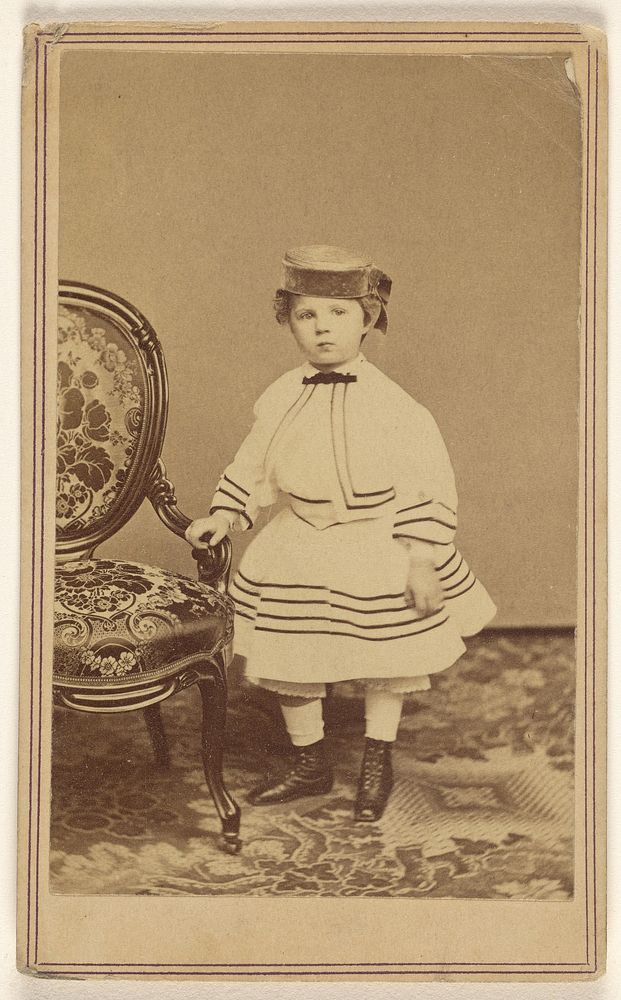 Unidentified little girl wearing a pill-box hat, standing, holding the arm of a chair by Churchill and Denison