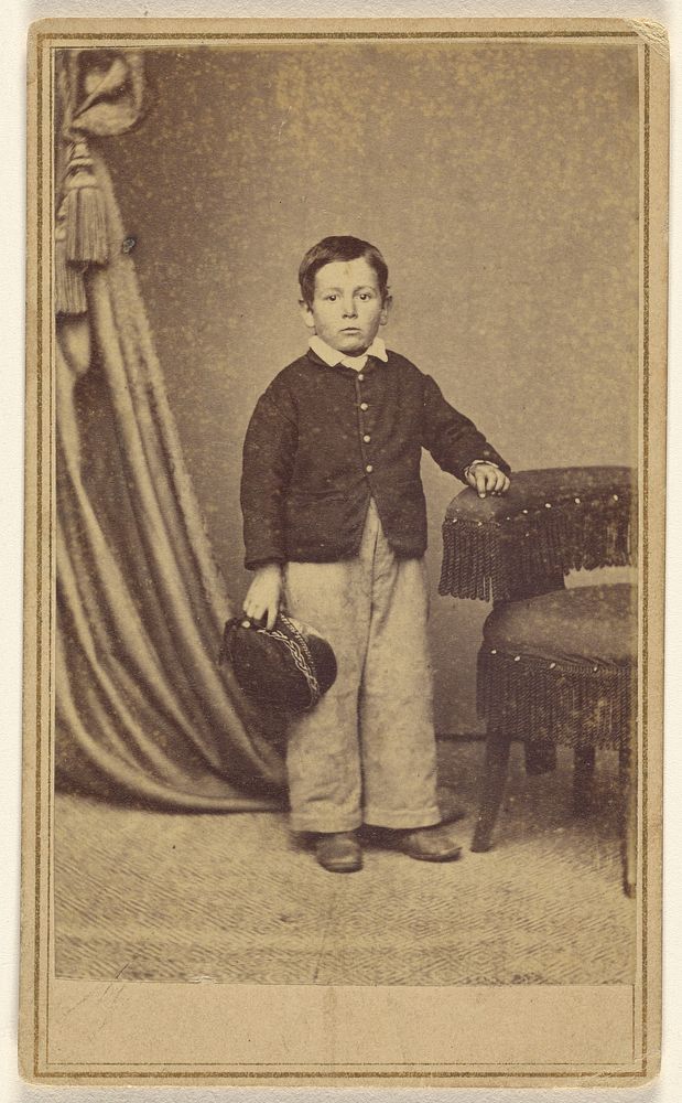 Unidentified little boy standing, holding his cap, with hand on chair back with tassels by H Jaeger and Company