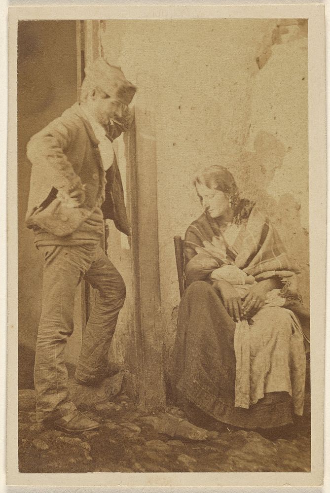 Man with round cap standing next to a seated woman with baby