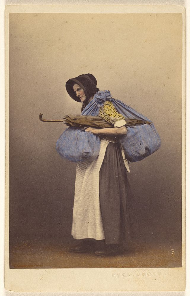 Unidentified elderly woman wearing a bonnet and native costume standing, carrying sacks and a parasol by Thomas Edge