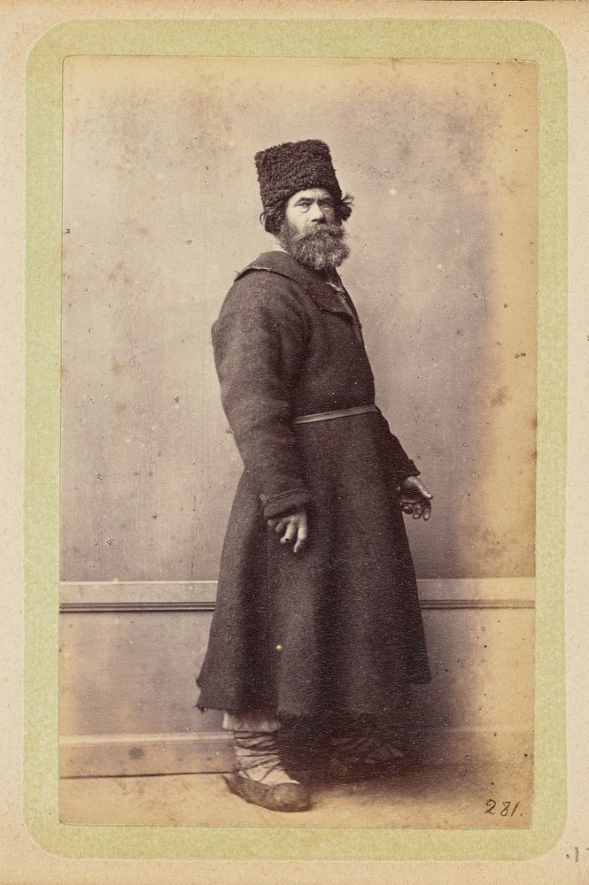 Bearded man posing in long, dark coat and wool hat by William Carrick