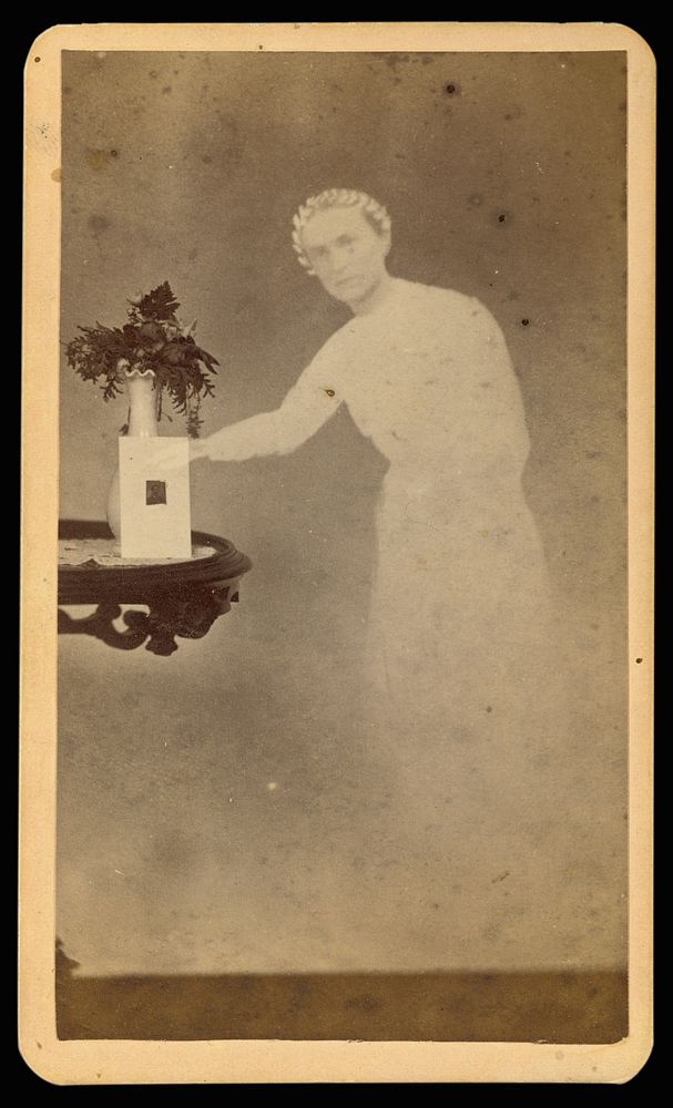 Female "spirit" standing next to a table with a photograph propped against a vase with flowers by William H Mumler and Helen…