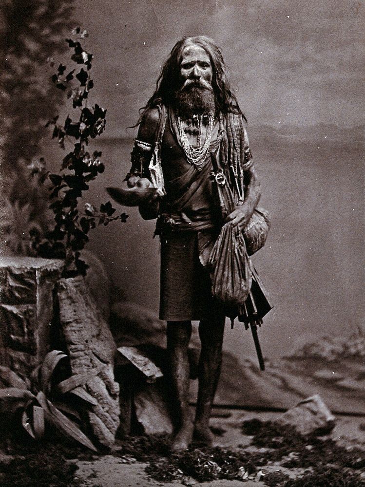 An Indian man wearing many beads, and carrying a begging bowl and an umbrella, in a studio setting. Photograph, ca.1900.