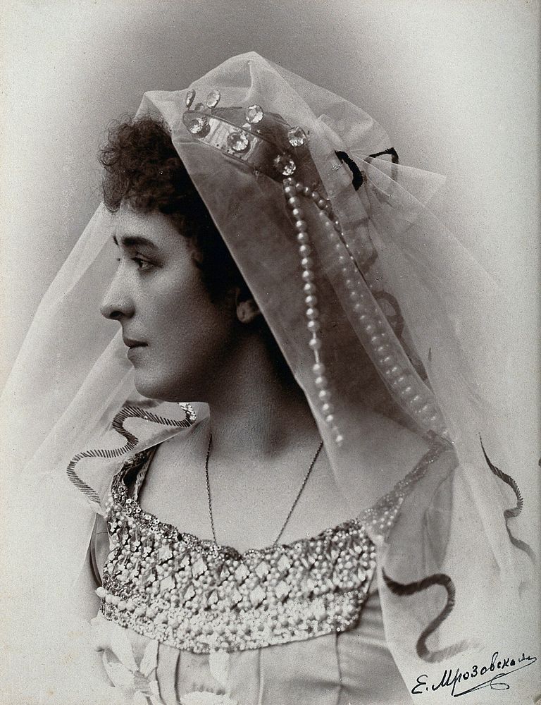 A woman posing in a photographic studio, wearing a bridal dress and veil, in front of a plain backdrop.