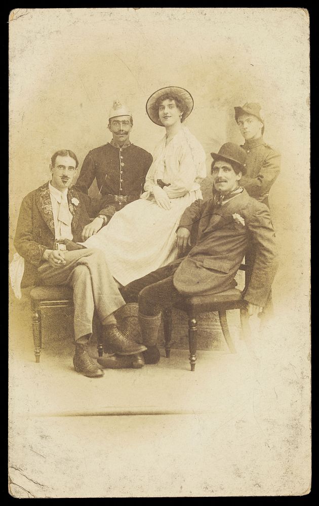 Amateur actors sitting down, surrounding a man in drag seated in the centre. Photographic postcard, 191-.