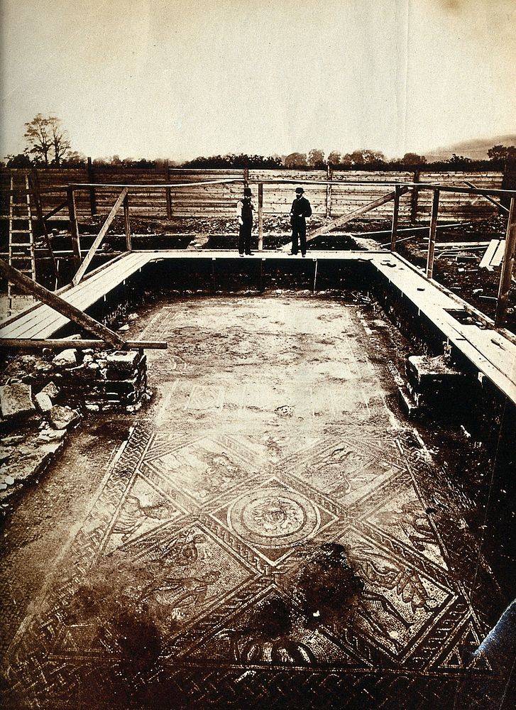 A Roman villa under excavation: two men stand on a platform looking at a mosaic floor. Photograph, 1860/1880.