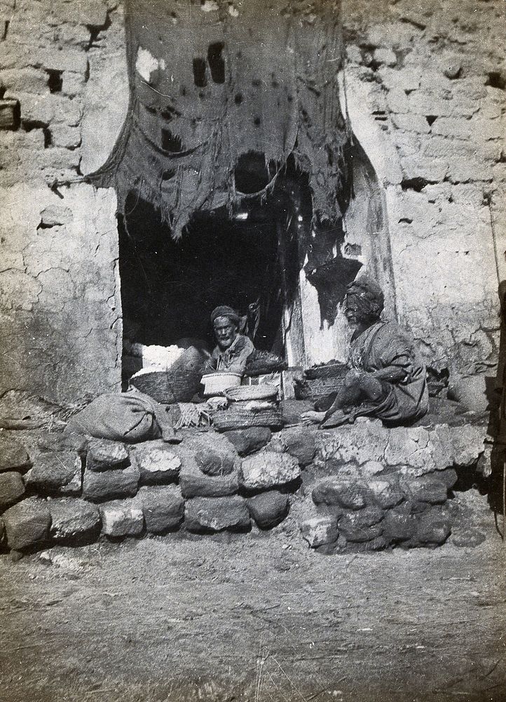 Shuster , Iran: doorway of a shop with two older men in turbans seated next to baskets of produce. Photograph, 1910/1920 .