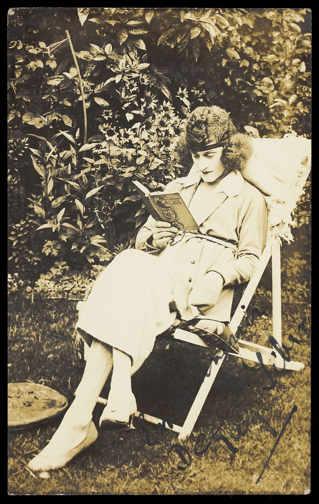 A sailor in drag, wearing a long coat and hat, sits reading in a garden. Photographic postcard, ca. 1920.