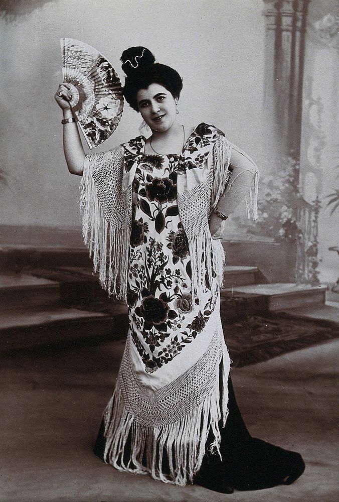 A standing woman, holding aloft a fan, wearing a fringed shawl, in front of a painted backdrop in a photographic studio.…